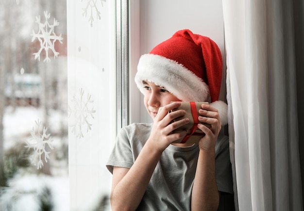 Um menino com um chapéu de Papai Noel abraça um presente e olha pela janela com ar sonhador