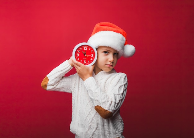 Um menino com um chapéu de Papai Noel segura um relógio em um fundo de estúdio vermelho o conceito de Natal e ano novo uma criança em roupas de malha