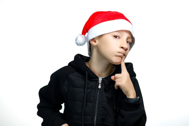 Um menino com um chapéu de Papai Noel vermelho sobre um fundo branco leva o dedo aos lábios e pensa