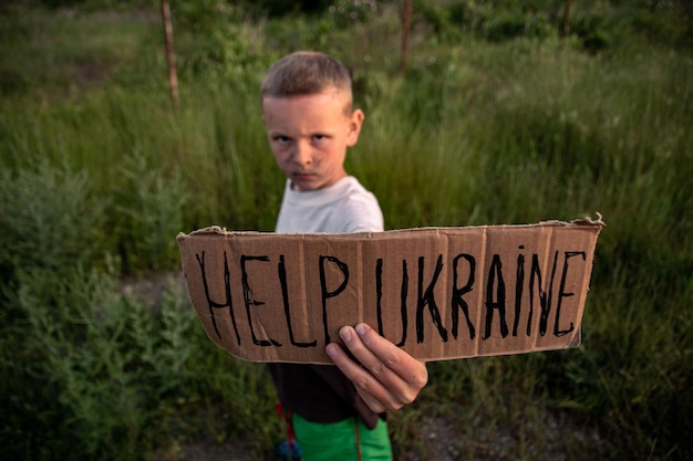 Um menino com um rosto sujo e olhos tristes segura um pôster de papelão com a inscrição HELP UKRAINE