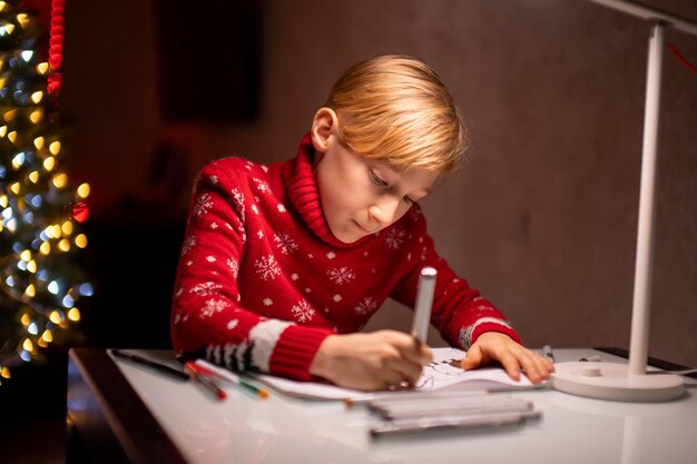 Um menino com um suéter vermelho de Natal no fundo de uma árvore de Natal desenha diligentemente atrás de uma lâmpada que brilha sobre ele