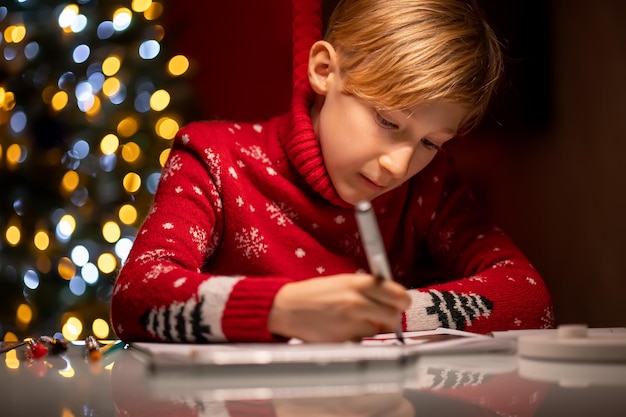 Um menino com um suéter vermelho de Natal no fundo de uma árvore de Natal segura um marcador para desenhar