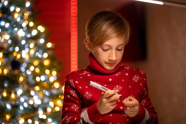 Um menino com um suéter vermelho de Natal no fundo de uma árvore de Natal segura um marcador para desenhar