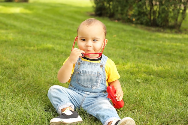 um menino com uma camiseta amarela está sentado no gramado do parque segurando um sino vermelho