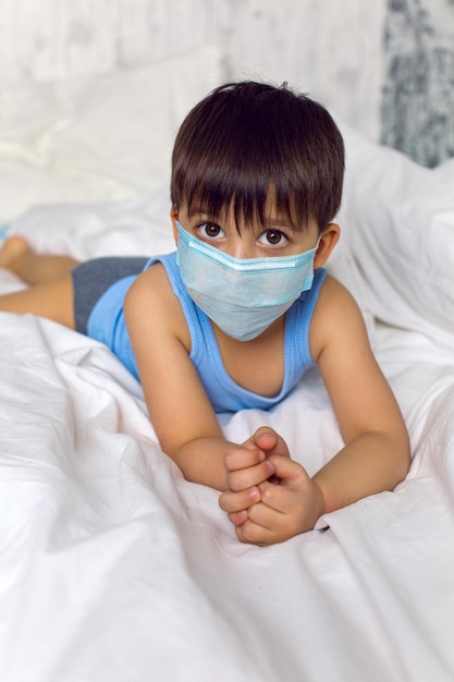 Foto um menino com uma camiseta azul e uma máscara médica senta-se em uma cama em quarentena durante a epidemia de covid-19