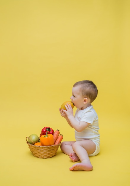 Foto um menino com uma cesta de legumes e frutas em um espaço amarelo. o bebê se senta de lado e come um limão. orientação vertical. copie o espaço