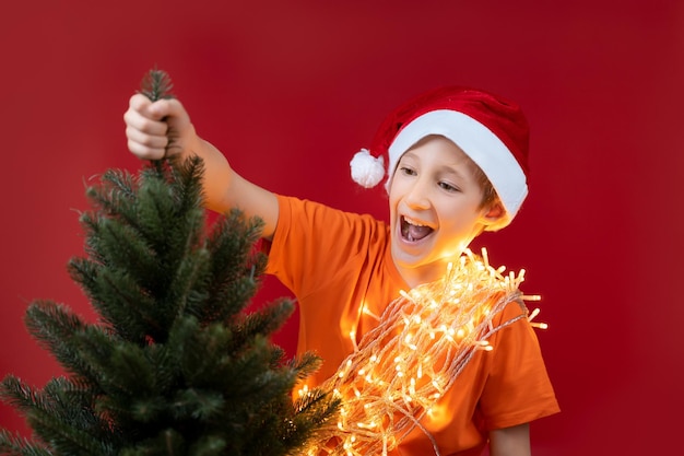 Um menino com uma guirlanda de Natal no pescoço e um chapéu de Papai Noel segura o topo de uma árvore de Natal e grita com ela