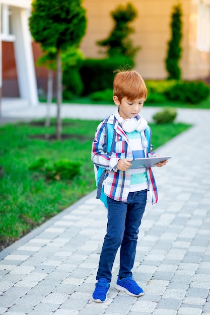 Um menino com uma mochila e um tablet com fones de ouvido na escola lendo um livro ou fazendo lição de casa o conceito está de volta à escola