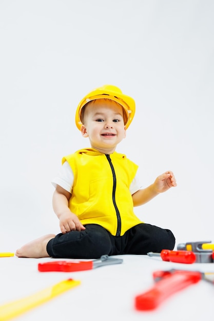Um menino de 2 anos de idade em um terno de construtor em um fundo branco Uma criança em um capacete de plástico e ferramentas de plástico Brinquedos infantis de plástico