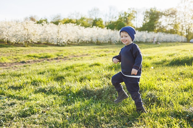Um menino de 3 anos com um suéter e um chapéu corre por um jardim florido Roupas para crianças de 3 anos Uma criança eotiana feliz é branca entre macieiras em flor