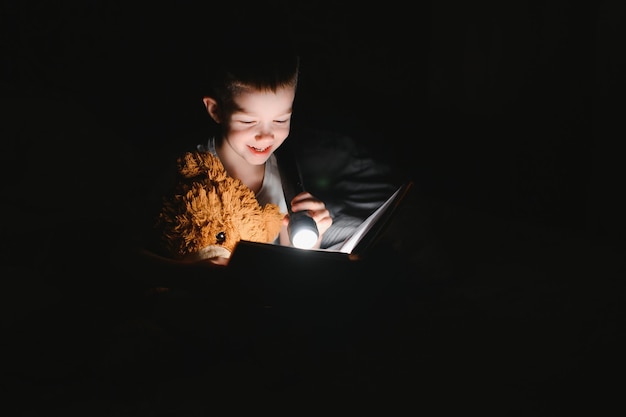 Um menino de 5-6 anos está lendo um livro à noite no escuro sob um cobertor com um urso de brinquedo