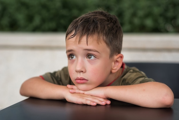 Um menino de 6 anos com uma camiseta com uma estampa do exército está sentado a uma mesa com a cabeça baixa nas mãos