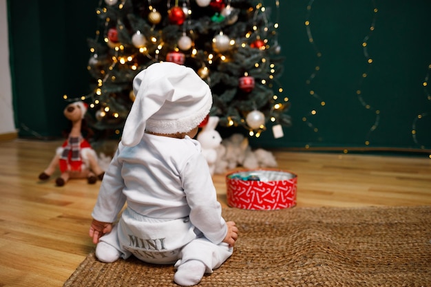 Um menino de 9 meses perto de uma árvore de Natal em um terno branco quente e chapéu Feliz Natal em casa feriado