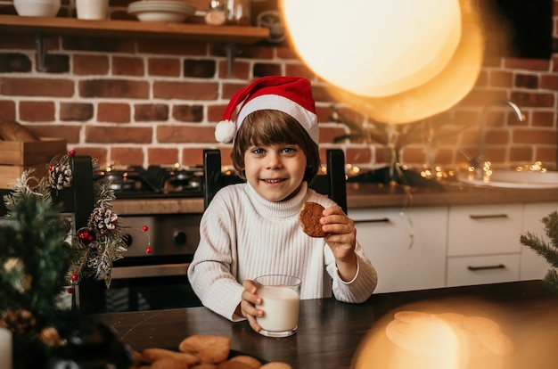 Um menino de ano novo está sentado à mesa da cozinha com um suéter branco e um chapéu vermelho de ano novo com leite e biscoitos