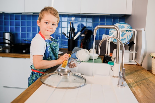 Foto um menino de avental lava pratos na cozinha de casa.