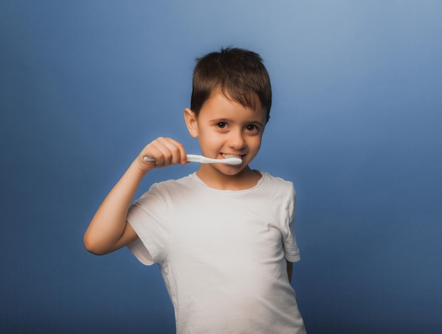 Um menino de cabelos escuros com uma camiseta branca sobre um fundo azul escova os dentes com uma escova de dentes. higiene do bebê