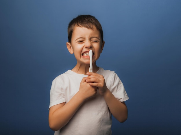 Um menino de cabelos escuros com uma camiseta branca sobre um fundo azul escova os dentes com uma escova de dentes. higiene do bebê