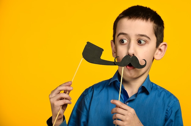 Um menino de camisa azul em um fundo amarelo faz uma careta e veste um bigode e segura um cachimbo