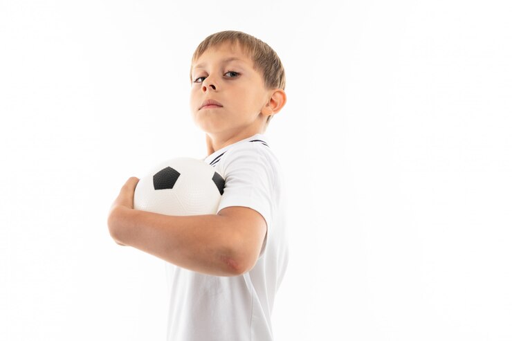 Foto um menino de camisa branca, calção azul com cabelos loiros, óculos escuros com bola de futebol