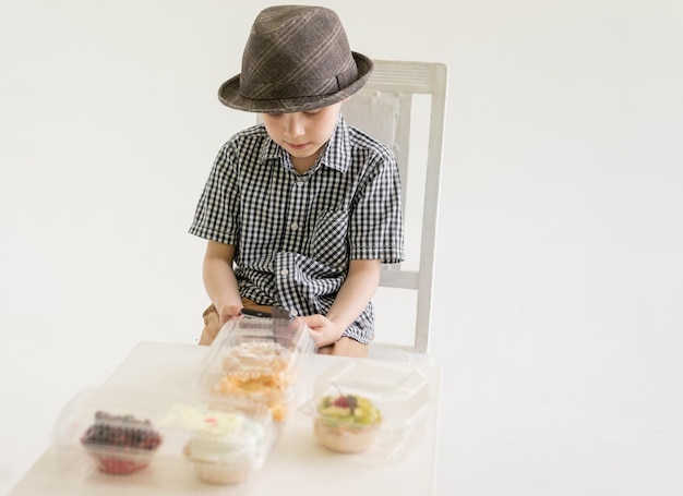 Um menino de camisa e chapéu se senta na frente de bolos com um telefone nas mãos