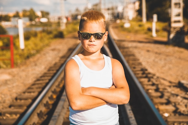 Um menino de camiseta branca e óculos pretos está parado na ferrovia.