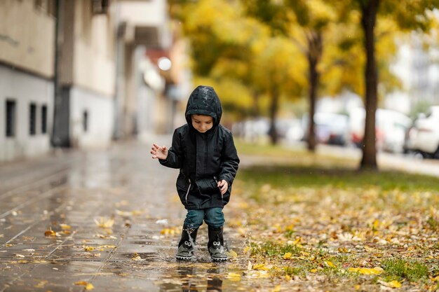 Um menino de capa de chuva e botas está brincando na rua e pulando na poça na rua