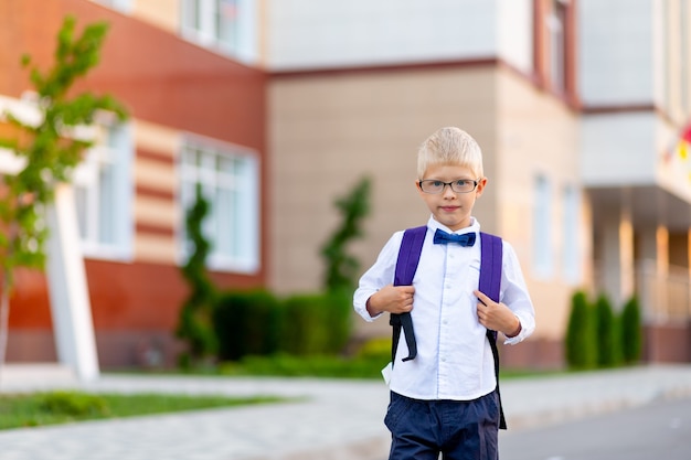 Um menino de escola com óculos louros e uma mochila está parado na escola