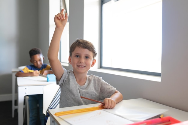 Um menino de escola primária caucasiano sorridente com a mão levantada sentado na mesa na sala de aula