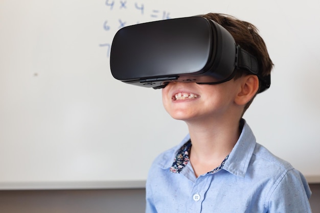 Um menino de escola primária caucasiano sorridente usando óculos VR contra o quadro branco na sala de aula