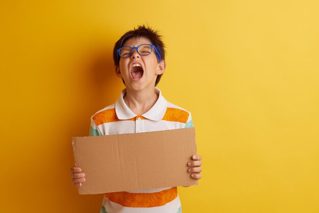 um menino de óculos em um fundo amarelo segura um banner de papelão com um espaço vazio para texto