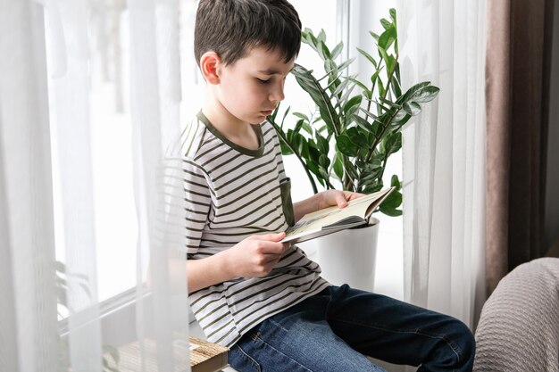 Um menino de seis anos está lendo um livro enquanto está sentado na janela Escola em casa