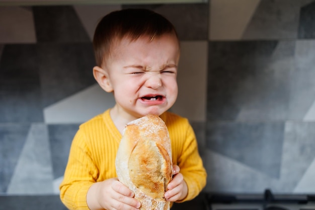 Um menino de um ano com roupas amarelas senta e come pão de centeio recém-assado A criança segura uma baguete fresca em suas mãos