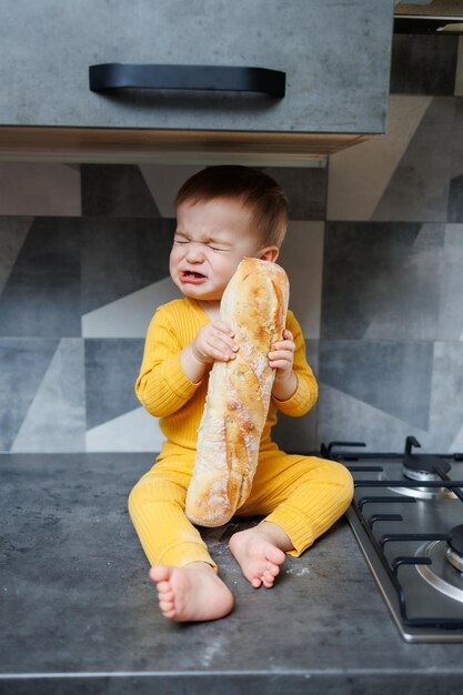 Um menino de um ano com roupas amarelas senta e come pão de centeio recém-assado A criança segura uma baguete fresca em suas mãos