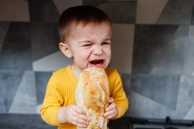 Um menino de um ano com roupas amarelas senta e come pão de centeio recém-assado A criança segura uma baguete fresca em suas mãos