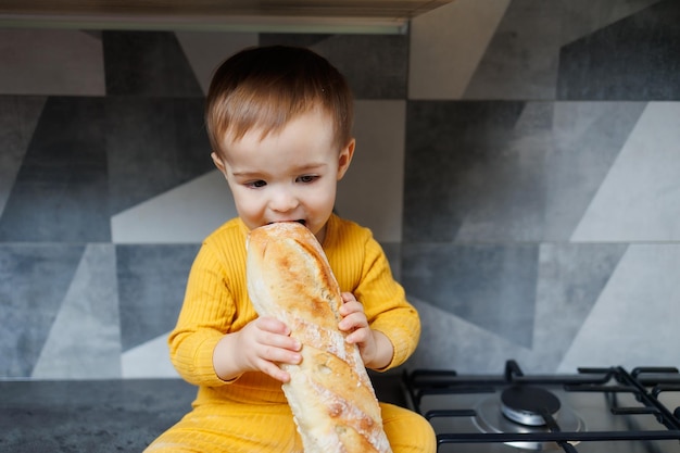 Um menino de um ano com roupas amarelas senta e come pão de centeio recém-assado A criança segura uma baguete fresca em suas mãos