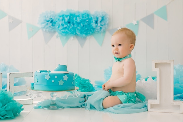 Um menino de um ano de bermuda azul e gravata borboleta comemora seu aniversário ao lado de um bolo