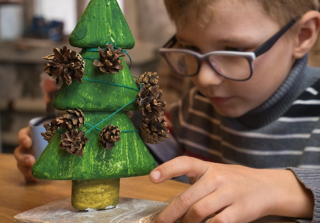 Um menino decora uma árvore de Natal feita à mão com pinhas e ao mesmo tempo brinca de forma engraçada.