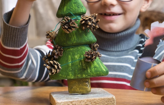 Um menino decora uma árvore de Natal feita à mão com pinhas e ao mesmo tempo brinca de forma engraçada.