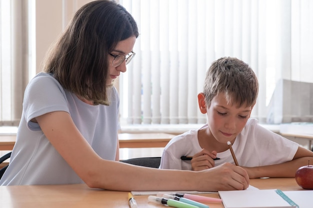 Um menino e um professor estão sentados em uma mesa com cadernos e canetas A mãe observa seu filho escrever em um caderno