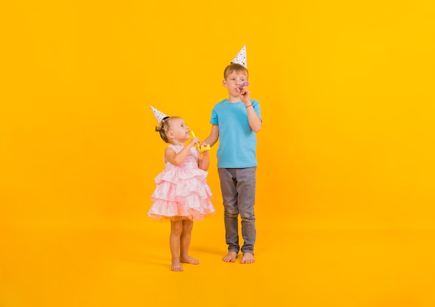 Um menino e uma menina se levantam e celebram a celebração em bonés e apitos de papel soprando em um fundo amarelo com uma cópia do espaço