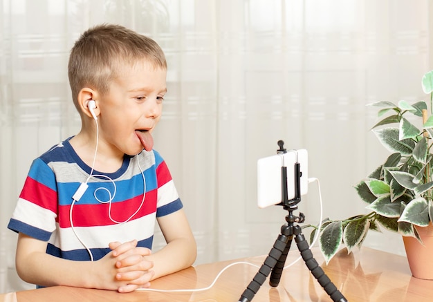 Um menino em fones de ouvido olha para o telefone mostrando a língua Vício infantil em gadgets Educação online para pré-escolares