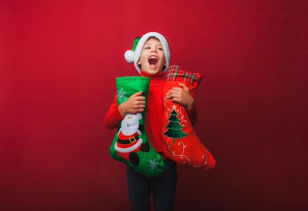 Um menino em um suéter vermelho de malha e um chapéu de papai noel segura uma bota de natal para presentes bebê fofo está esperando o natal e um novo tipo de isolamento em um fundo vermelho