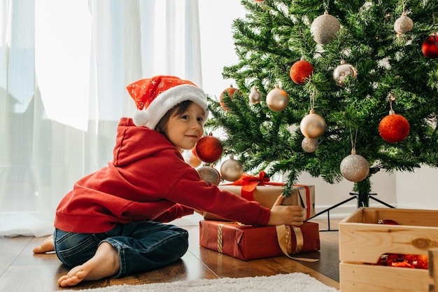 Um menino encantador com um chapéu de Papai Noel tira presentes de Natal de debaixo da árvore de ano novo e ri