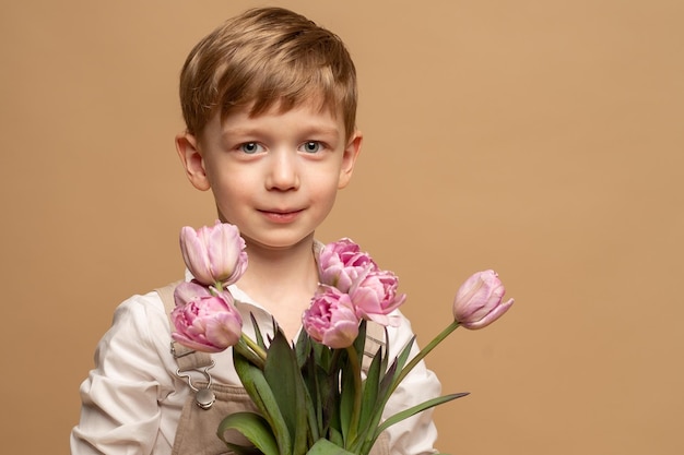 um menino encantador de quatro anos de macacão bege e uma camisa branca segura um buquê de tulipas cor de rosa