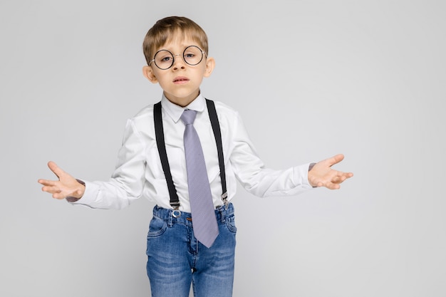 Um menino encantador em uma camisa branca, suspensórios, uma gravata e jeans leves fica em cinza