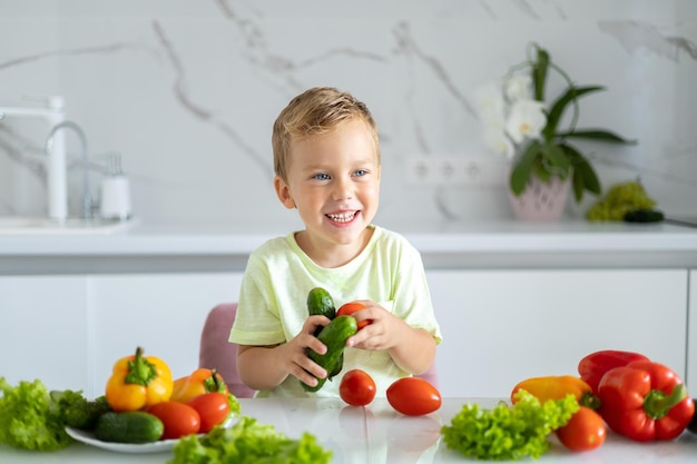 Um menino está sentado em uma mesa com legumes na cozinha em casa segurando pimenta nas mãos o bebê está sorrindo o conceito de alimentação saudável vegetariana