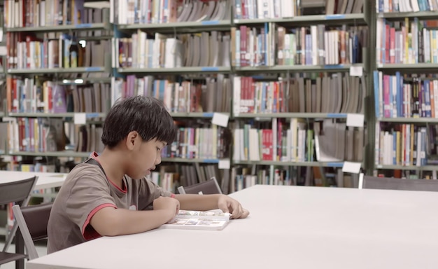 Um menino está sentado em uma mesa em uma biblioteca lendo um livro.