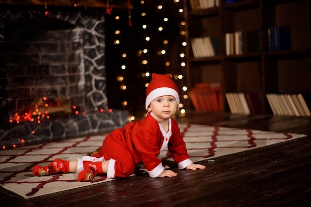 Um menino fantasiado de papai noel em um quarto decorado de natal perto da árvore de natal em um fundo de luzes.