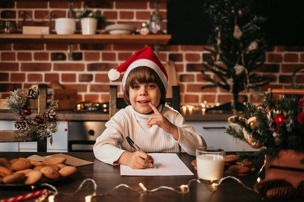 Um menino feliz com um suéter de tricô branco e um chapéu vermelho está sentado a uma mesa e escrevendo uma carta de Ano-Novo