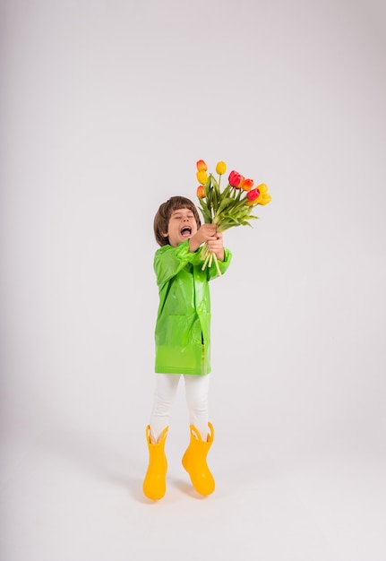 Um menino feliz com uma capa de chuva verde e botas de borracha amarelas segura um buquê de tulipas coloridas em um fundo branco com um lugar para texto